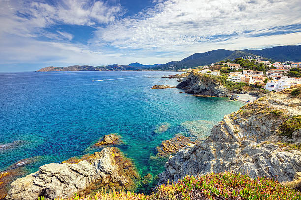 A coastal landscape features clear turquoise waters gently lapping against rocky shores. A village can be seen in the distance, nestled among the hills, with charming houses dotting the coastline. The sky is vast, filled with clouds that create a dynamic backdrop to the serene scene.