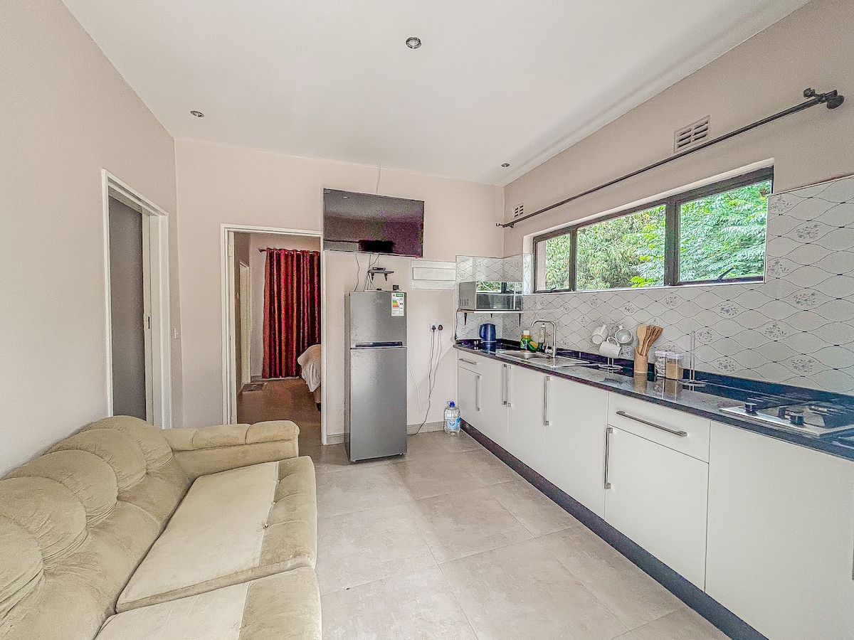 A functional kitchen is equipped with modern appliances, including a stainless steel refrigerator and stovetop. The space features light-colored cabinets and a countertop, complemented by large windows allowing natural light to fill the area. A comfortable beige couch offers a casual seating option.