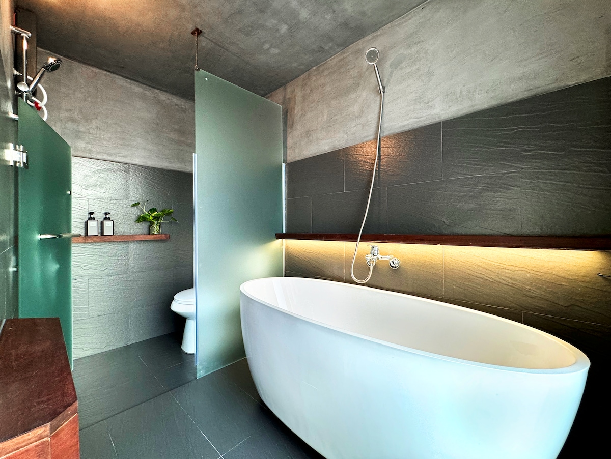 A modern bathroom features a sleek, freestanding bathtub and a glass shower enclosure. Minimalist wooden shelves hold toiletries and a small plant, contributing to the calming ambiance. Soft lighting accentuates the textured walls, enhancing the contemporary design.