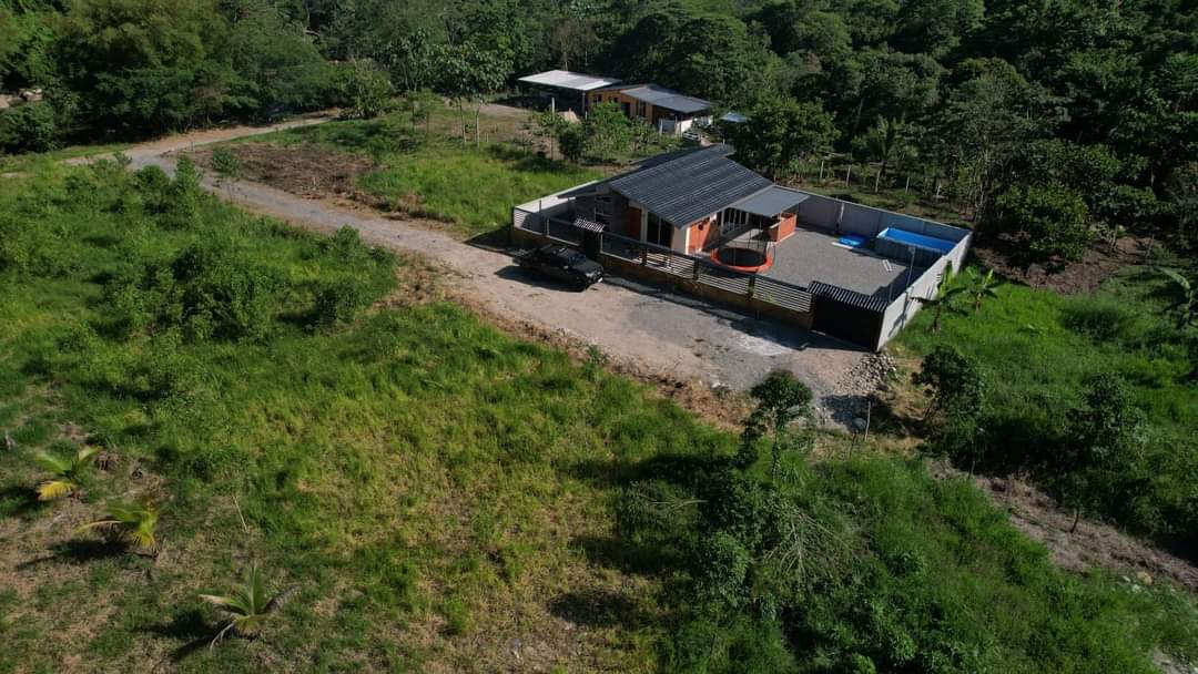 An aerial view captures a serene property surrounded by lush greenery. A comfortable home is positioned on a spacious lot, featuring an outdoor area with a blue swimming pool and a gravel driveway accommodating a parked car.