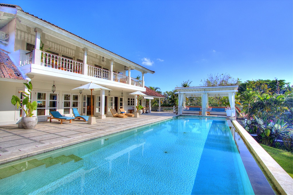 A spacious outdoor area features a large swimming pool, surrounded by a tiled deck with lounge chairs. The two-story villa is visible in the background, showcasing a blend of balconies and open spaces. Lush greenery enhances the tranquil atmosphere.