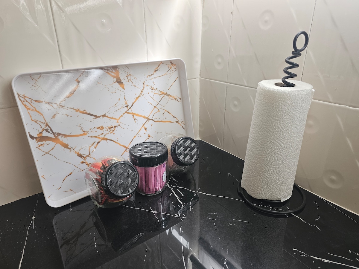 A marble-patterned tray is positioned on a sleek black countertop, accompanied by three jars in various colors. A spiral paper towel holder with a roll of paper towels sits nearby, creating a functional kitchen space.