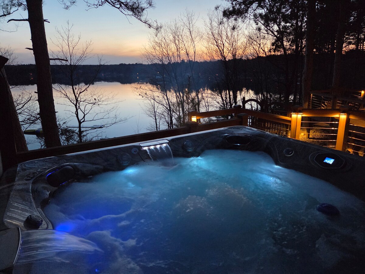 A serene hot tub is situated on the deck overlooking the lake, softly illuminated with blue lights. The tranquil water reflects the colors of the sunset, surrounded by trees, creating a peaceful atmosphere ideal for relaxation.