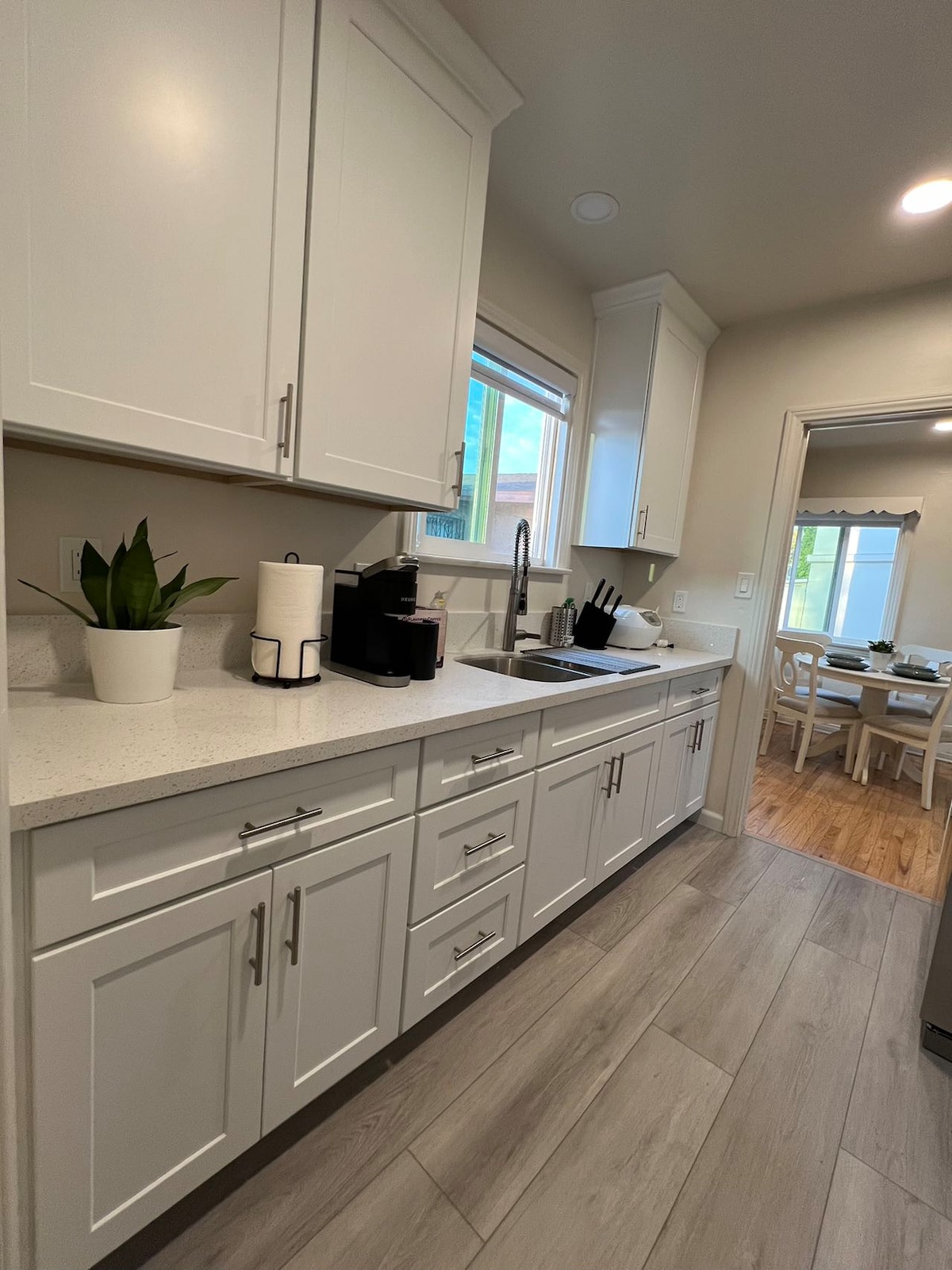 The kitchen features modern white cabinetry and a light countertop, providing ample storage. A coffee maker is set on the counter, near a plant and a sink. Natural light enters through the window, illuminating the space. The adjacent dining area is partially visible.