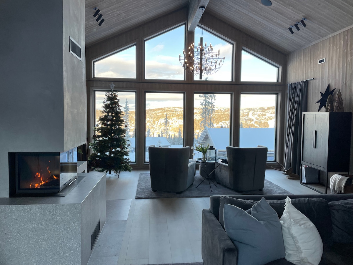 A spacious living area features large windows offering panoramic views of the surrounding mountains. Modern furniture includes two armchairs and a sectional sofa, while a serene fireplace adds a focal point. A Christmas tree is positioned nearby, contributing to the inviting atmosphere.