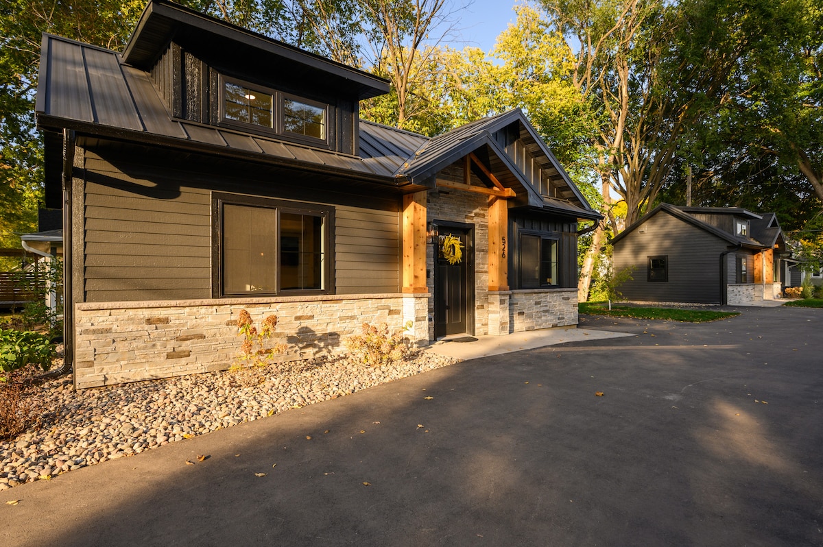 Two charming cottages are visible with modern exteriors, featuring a combination of dark siding and stone accents. The path in front is paved, with landscaped areas showcasing greenery. Large windows allow natural light to fill the interiors, while trees provide a serene backdrop.