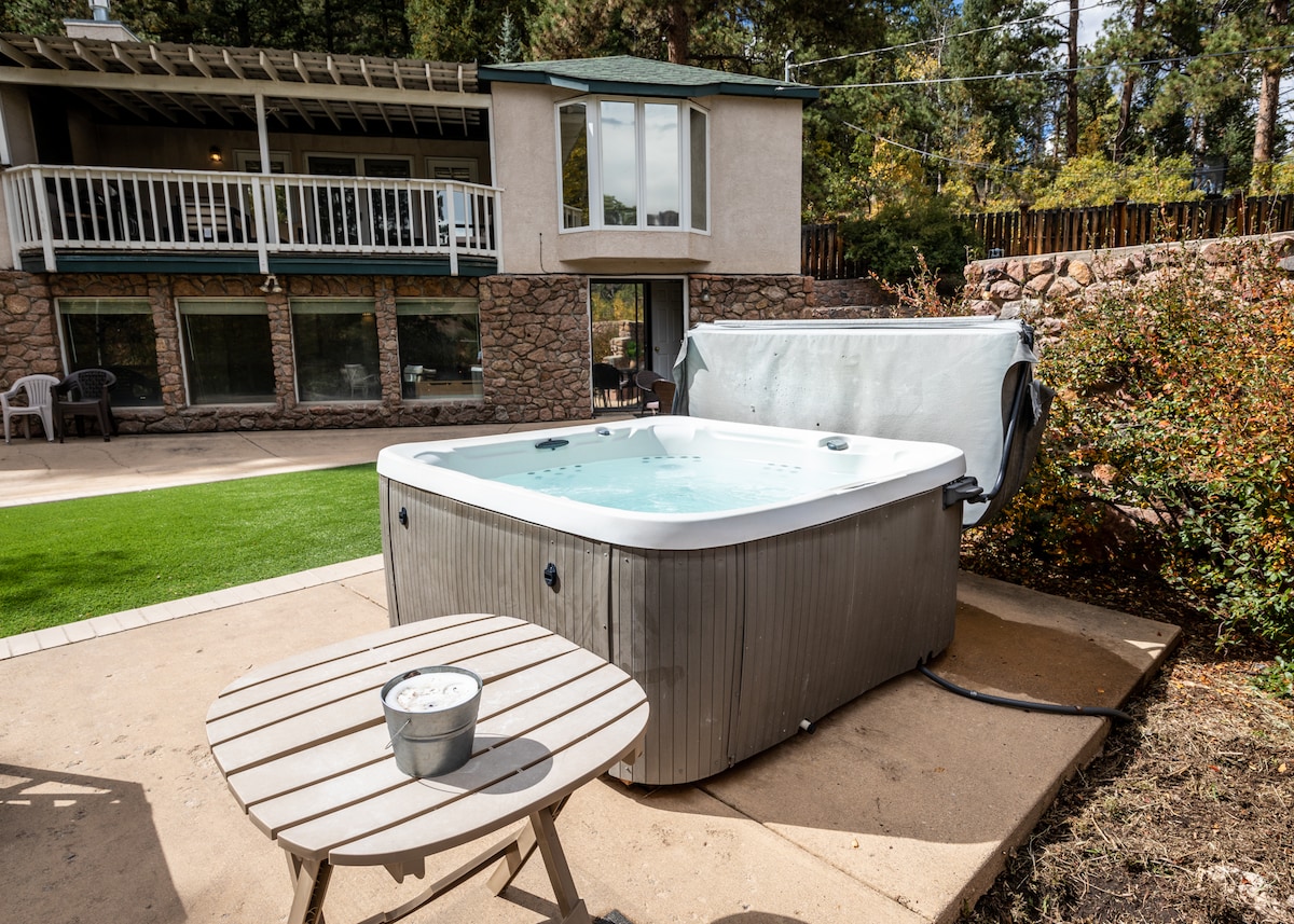 A hot tub sits on a stone patio, surrounded by lush greenery and a manicured lawn. Nearby, a small wooden table is positioned with a bucket on top, offering a convenient spot for refreshments. The backdrop features a multi-level home with an inviting outdoor space.