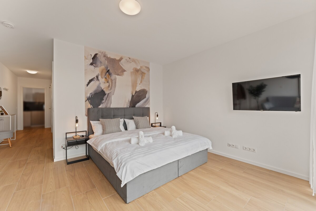 A spacious sleeping area features a contemporary box spring double bed, complemented by neutral bedding. A wall-mounted Smart TV is positioned above the bed, and soft lighting fixtures are placed on either side. Light wooden flooring and a decorative wall art piece enhance the modern design.