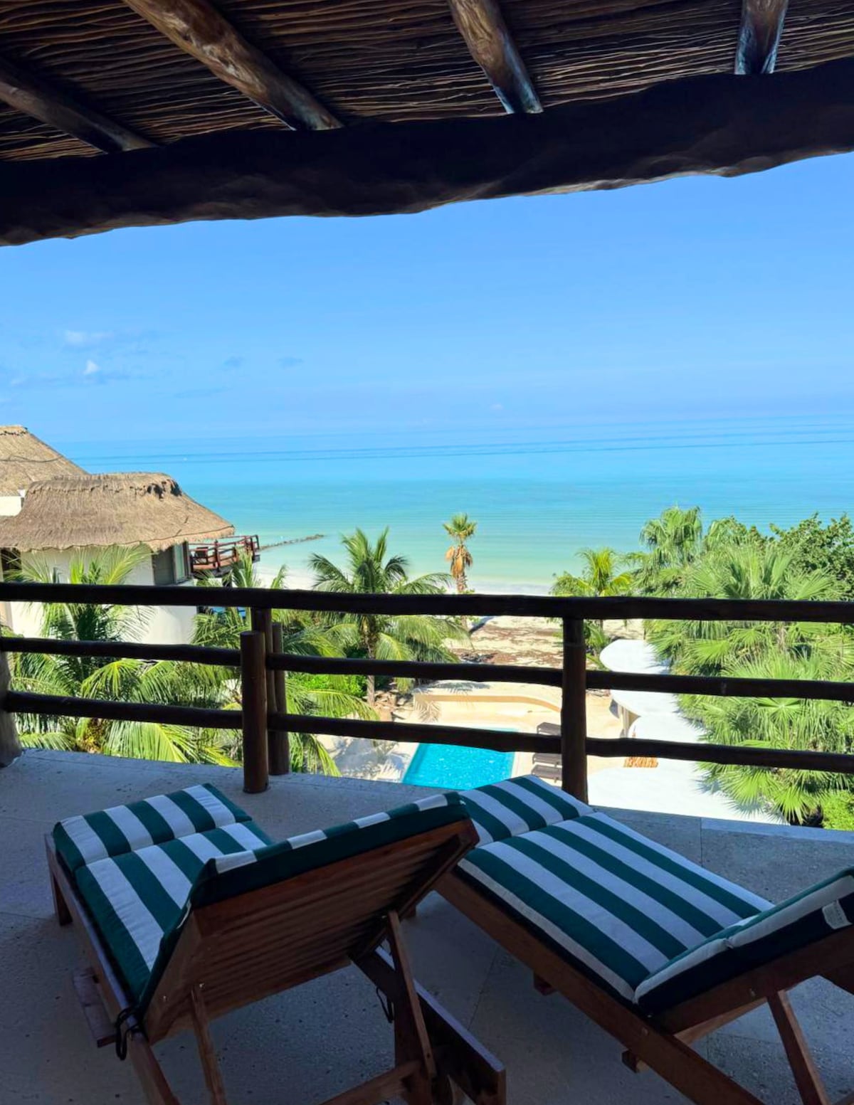 A serene view is captured from a private terrace, displaying two striped lounge chairs. The turquoise ocean stretches in the background, with lush palm trees framing the scene and a glimpse of a tranquil pool visible below.
