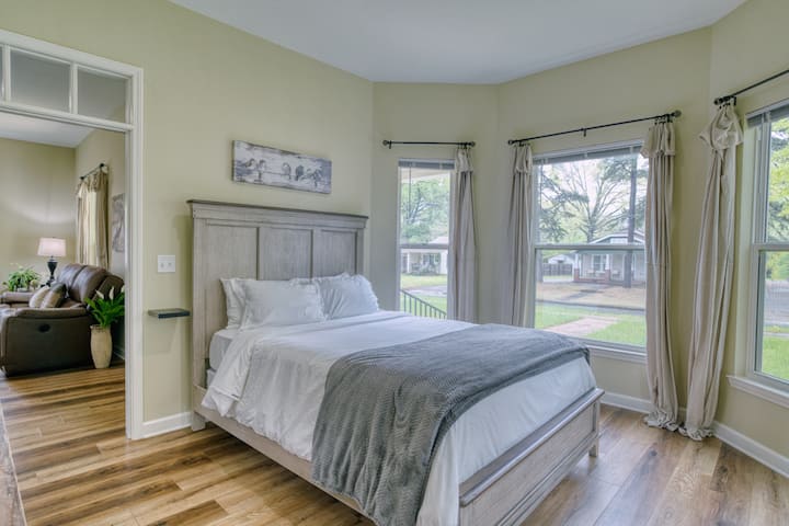 חדר שינה מואר עם מיטת קווין סייז הפונה לחלון מפרץ, ממוסגר על ידי וילונות בצבע בז' ונוף לחצר. כולל שמיכה נעימה, יצירות אומנות על הקיר בנושא ציפורים ודלת שמובילה לסלון. קירות ירוקים רכים ורצפות עץ.