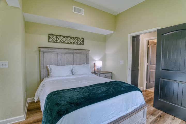 חדר שינה פשוט ונוח עם מיטת קווין סייז, ראש מיטה מעץ ותאורה רכה. יש שתי דלתות ורצפות מעץ, עם תליון קיר בסגנון בוהו מעל המיטה.