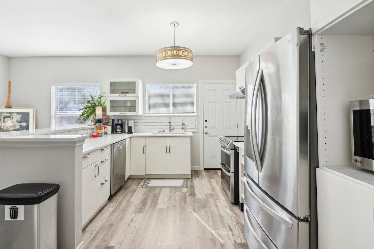 A modern kitchen features stainless steel appliances, including a refrigerator and stove. Clean lines are complemented by pale cabinetry and natural light from two windows. A circular pendant light hangs above a center island, enhancing workspace convenience. A door leads to the outdoor area.