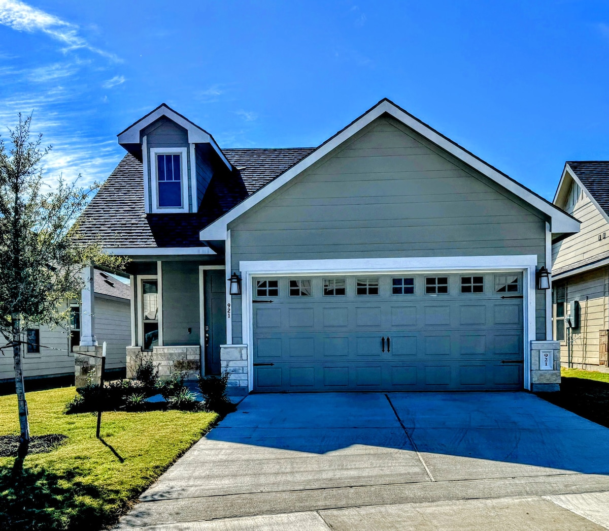 A modern, single-story house is featured with a light green exterior and a welcoming front porch. The double garage door is prominently displayed, surrounded by well-maintained landscaping that includes small shrubs and a neat lawn. The clear blue sky above adds to the inviting appearance.
