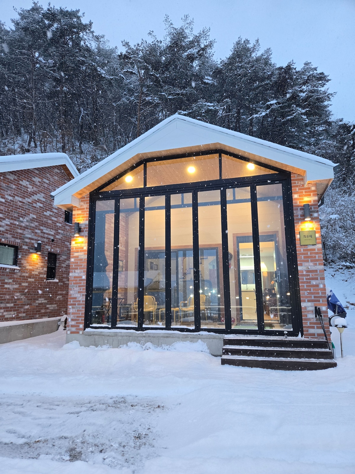The exterior of the house features large glass panels that allow natural light to fill the interior. Snow gently falls on the white and red-brick facade, accentuating the modern architecture. Soft lighting highlights the entrance, creating an inviting atmosphere in the snowy setting.
