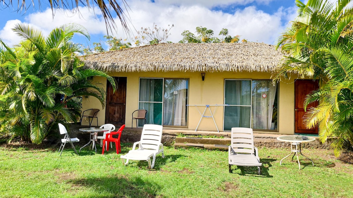 A rustic cabin with a thatched roof is set amidst lush greenery. Two wooden doors provide access to the interior, while large windows allow natural light to enter. Outdoor seating includes two white lounge chairs, a small table, and a red chair on the grass.