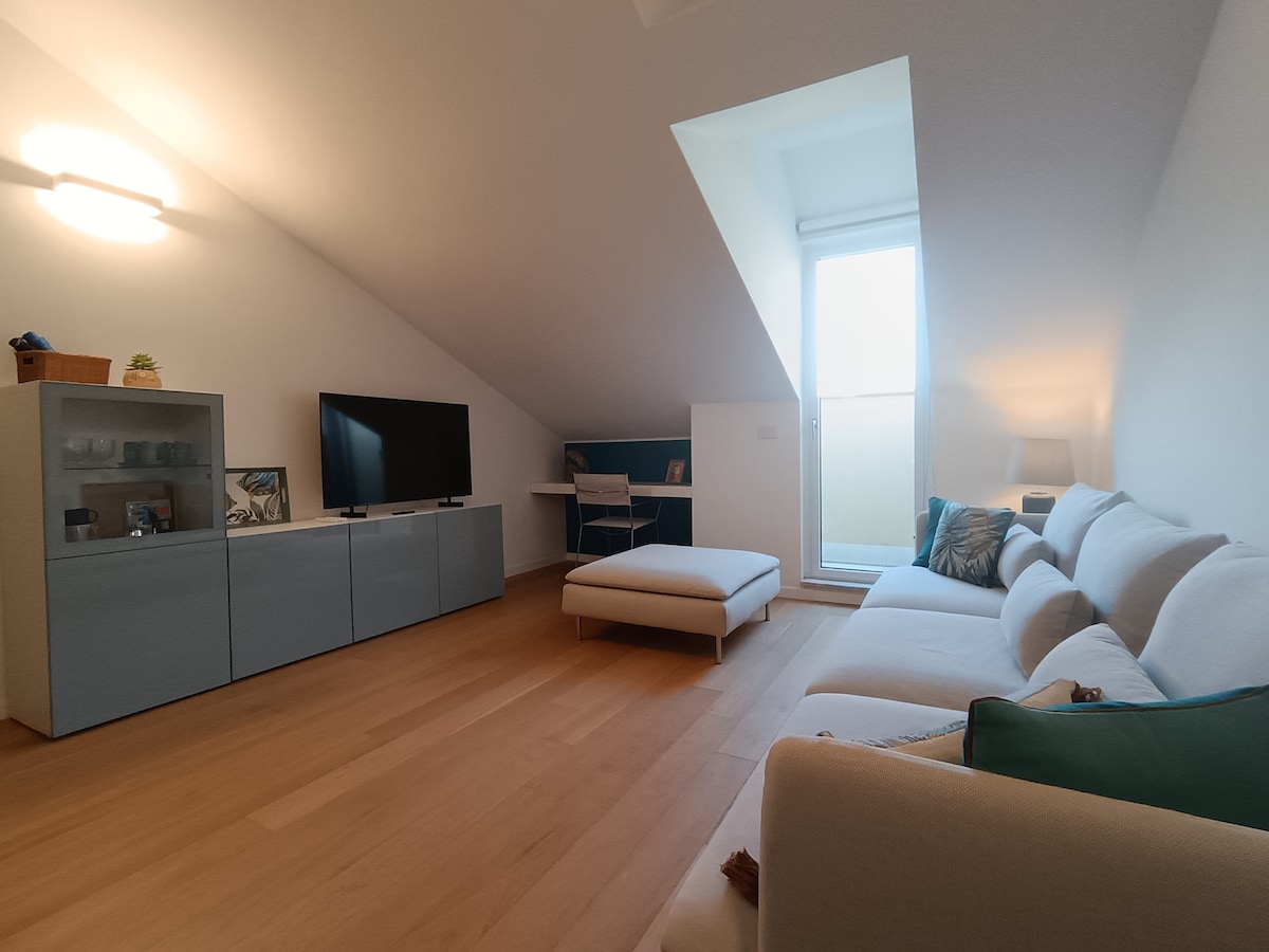 A spacious living area features a light-colored couch complemented by decorative pillows. A flat-screen television is positioned against a wall cabinet. Natural light enters through a large window, illuminating a small workspace with a desk and chair in a corner.