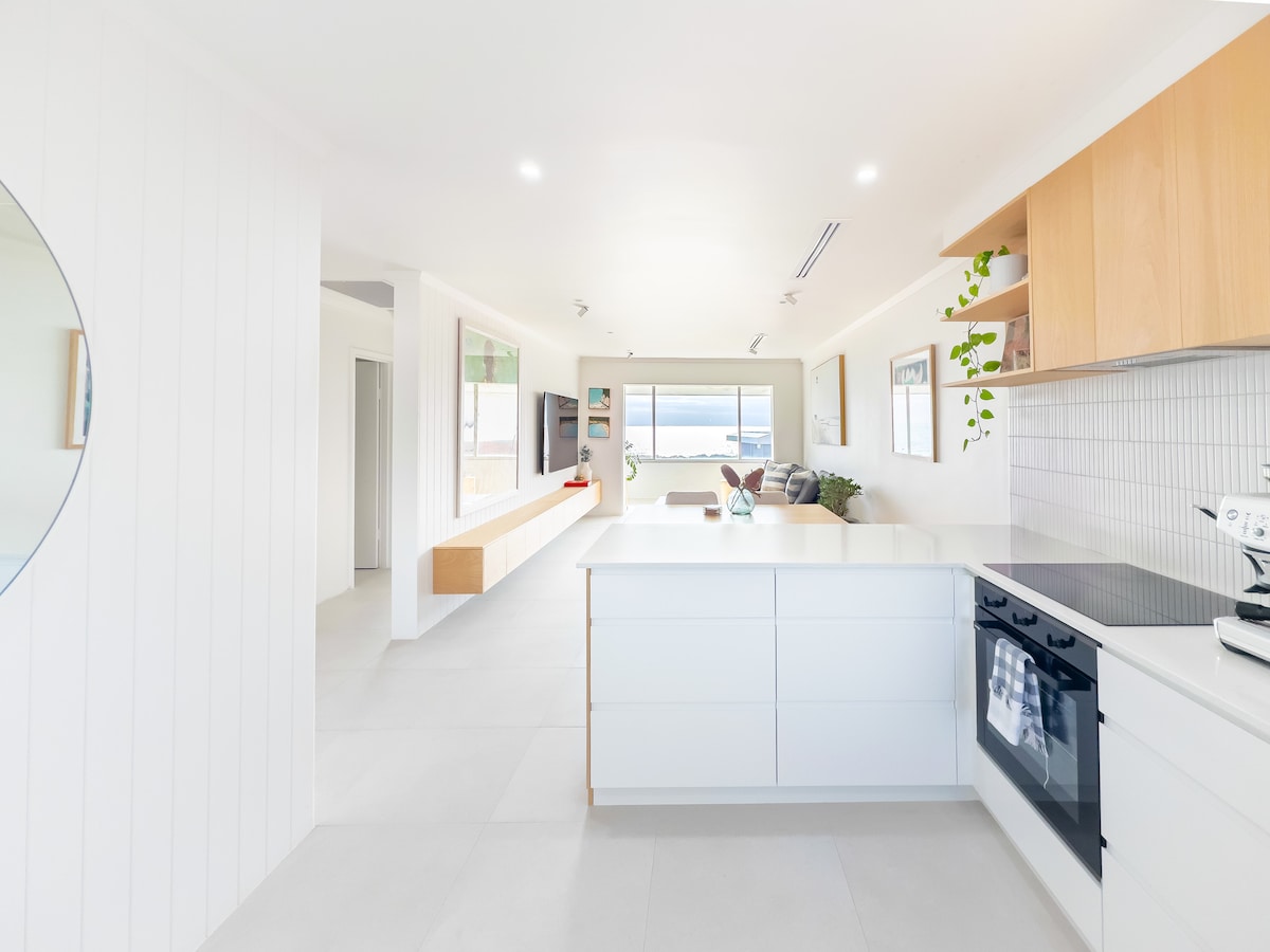 A modern kitchen is presented with sleek white cabinetry and a light wooden countertop. Natural light floods the space, which includes a stove and an oven. A round mirror reflects the open layout leading to a living area with ocean views.