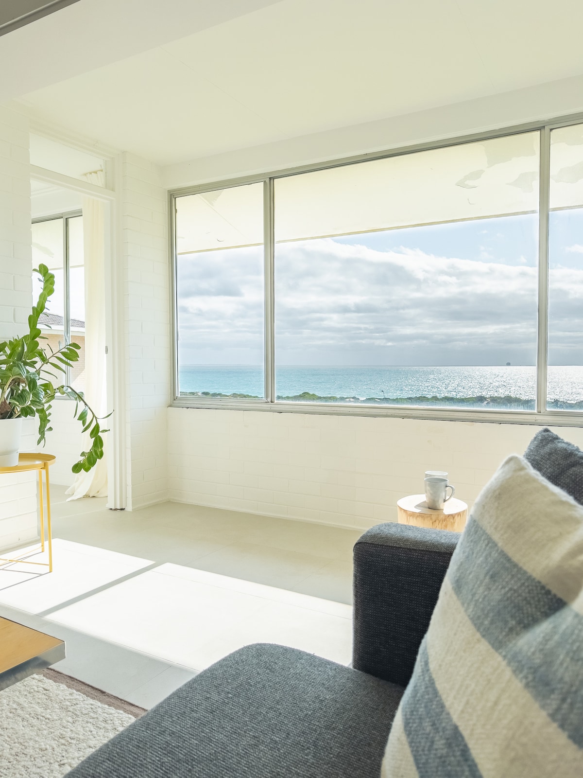 A bright living area features large glass windows offering expansive ocean views. The space is furnished with a dark sofa and a small side table, enhancing a serene coastal atmosphere. Greenery from a potted plant adds a touch of nature to the interior.