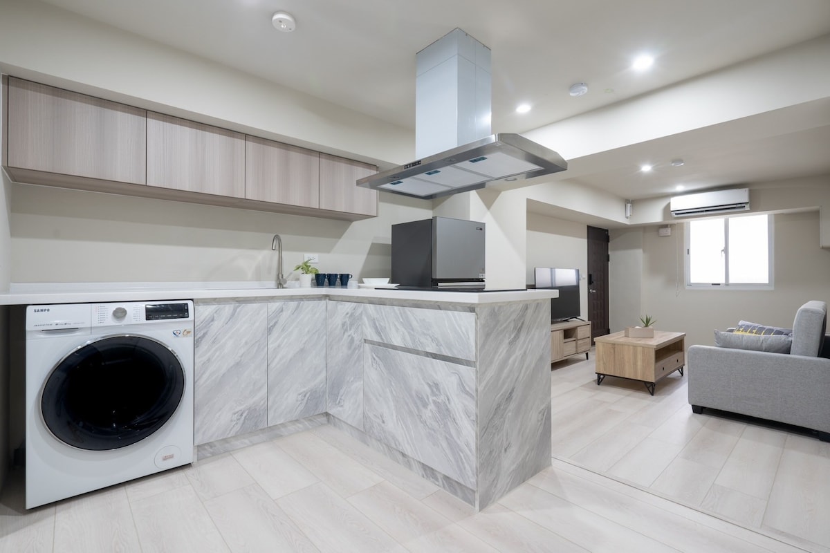 A modern kitchen area features sleek cabinetry and a marble countertop. Appliances include a washing machine and a stovetop, with a flat-screen television placed nearby. Natural light enters through a window, highlighting the light wooden flooring and inviting living space.