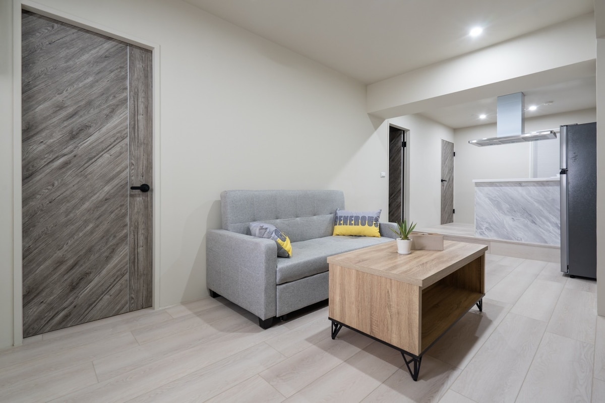 A modern living area features a light gray sofa beside a wooden coffee table. The room is illuminated by soft lighting, showcasing light-colored flooring and neutral walls. A door with a unique wood grain design is visible in the background, along with a minimal kitchen area.