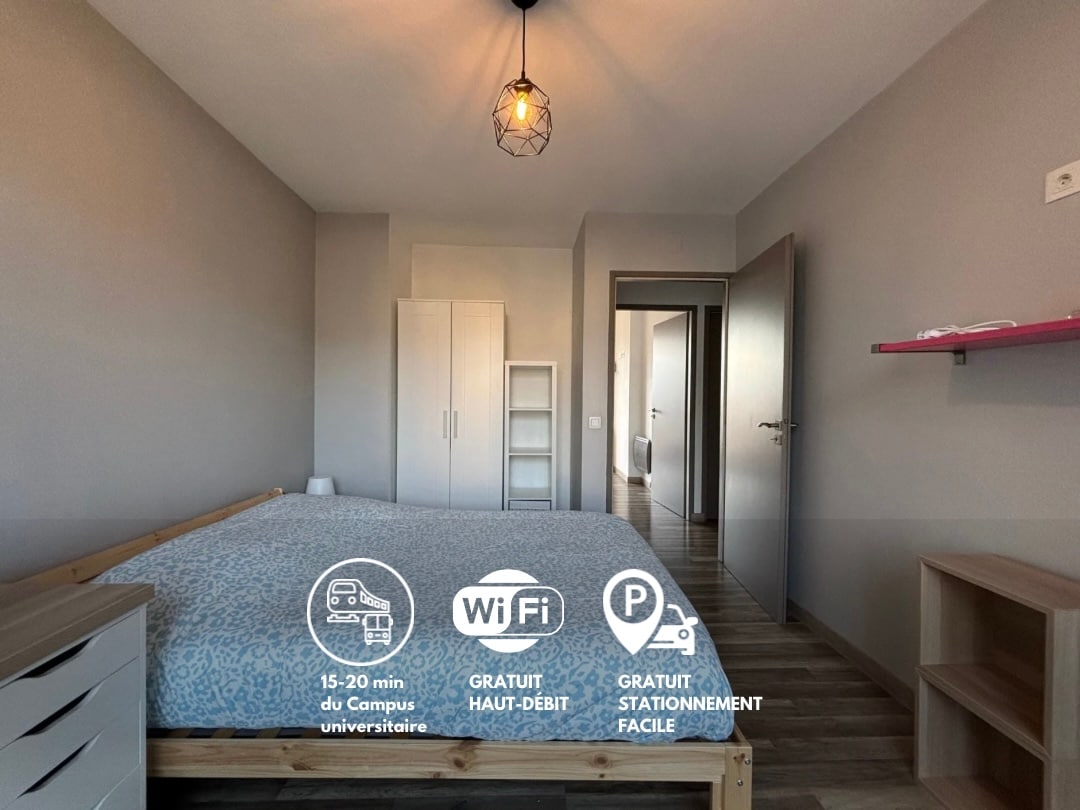 A spacious bedroom features a wooden bed adorned with a patterned duvet. A closet stands against the wall, complemented by a shelf displaying decorative elements. Natural light enters through the doorway, illuminating the neutral-colored walls and wooden floor.