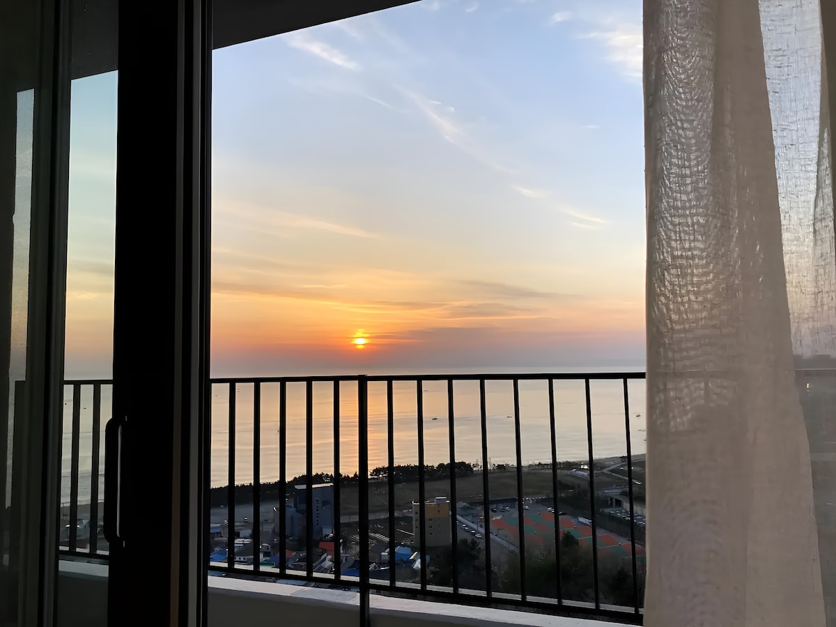 A serene view of the ocean is framed by an open balcony door. The sunset casts vibrant hues across the sky, reflecting on the calm waters. Gentle waves can be seen in the distance, complemented by a light texture from the sheer curtain softly swaying.