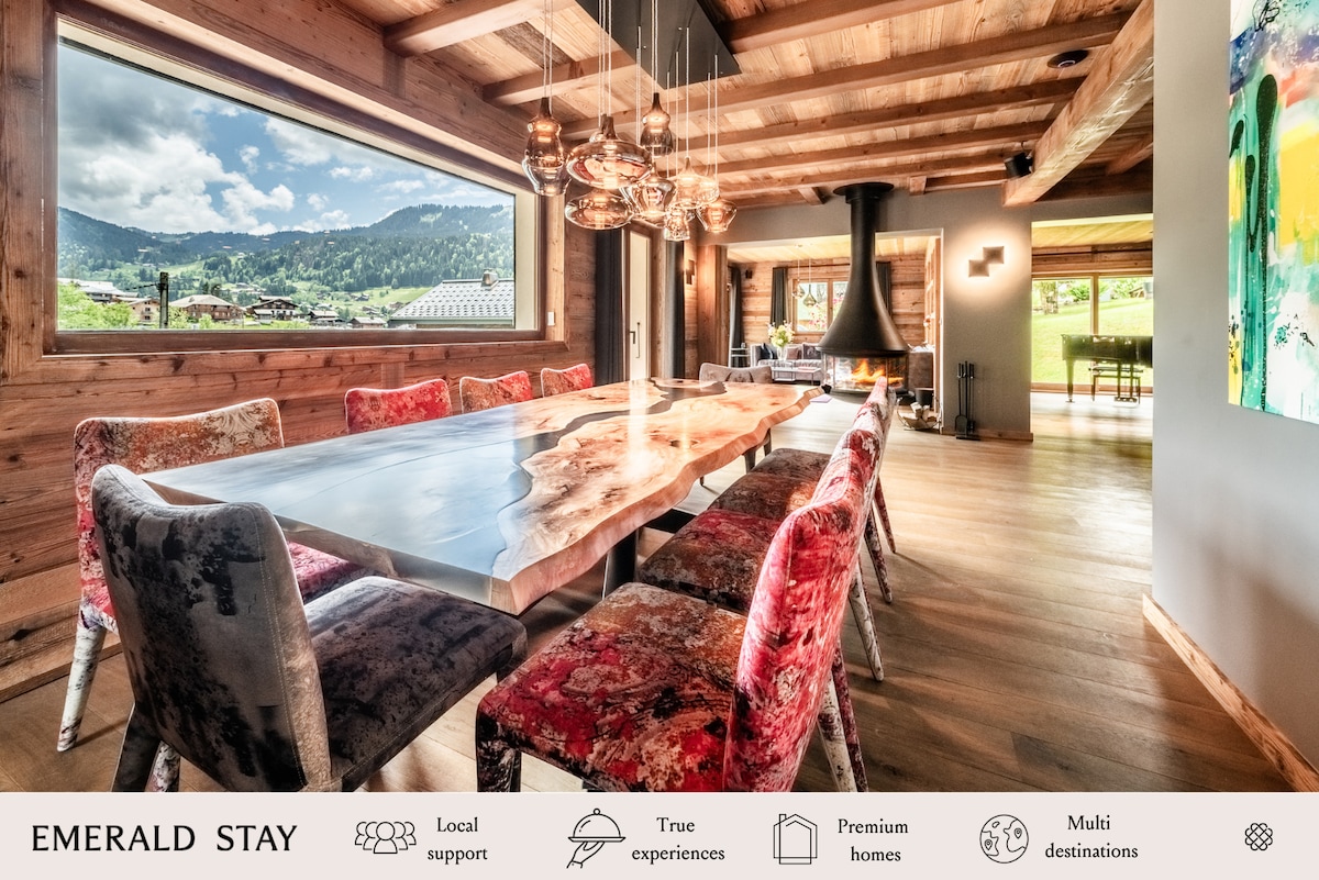 A spacious dining area is highlighted by a large wooden table with a live edge surface, surrounded by colorful upholstered chairs. Large windows frame mountain views while natural light fills the room, accentuating the rustic wooden beams and contemporary lighting fixtures above.