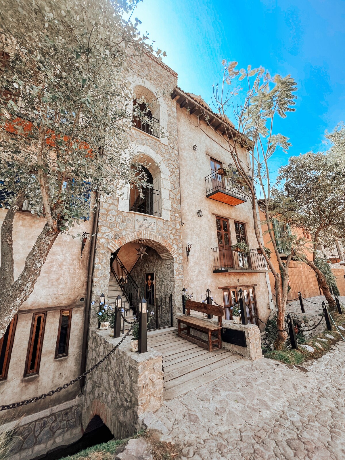 The exterior of a charming stone building is visible, featuring arched doorways and wooden balconies. Lush trees frame the scene, while a cobblestone pathway leads to the entrance. A rustic wooden bench is positioned on the porch, inviting guests to relax and enjoy the surroundings.