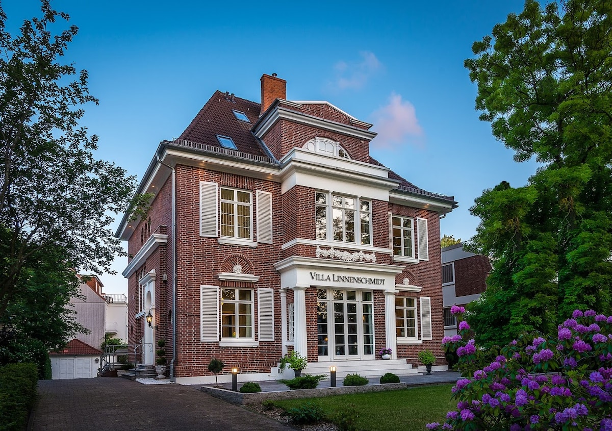 The elegant brick exterior of the villa is highlighted by large windows and classic architectural details. Lush greenery surrounds the entrance, while vibrant flowers add color to the well-maintained garden. The tranquil scene is complemented by a clear evening sky.