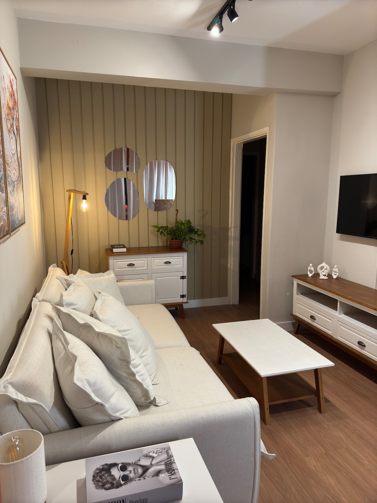 A cozy living area features a light-colored sofa adorned with four decorative pillows. A small coffee table sits in front of the sofa, and a stylish side table holds a lamp. A wall-mounted TV and decorative mirrors enhance the space, complemented by a small potted plant.