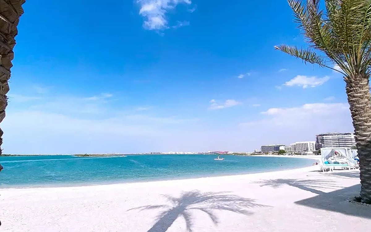 A serene beach view features soft white sand meeting calm turquoise waters under a clear blue sky. Palm trees cast gentle shadows on the shore, while distant buildings line the horizon, offering a glimpse of the coastal landscape.