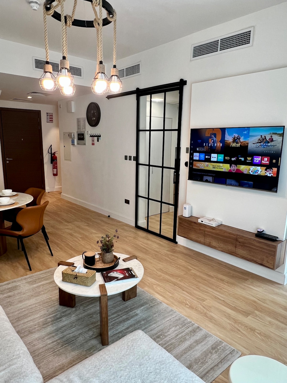 A cozy living area features a mounted smart TV displaying streaming options. A round coffee table with decorative items is centered on a light rug. The space is illuminated by an industrial-style pendant light and features warm wood accents throughout.