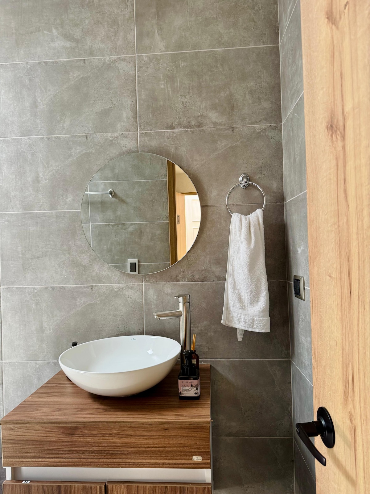 A modern bathroom features a circular mirror above a contemporary vessel sink. Soft natural light highlights the neutral-toned wall tiles, while a neatly hung towel is displayed on a ring beside the sink. The wooden cabinetry adds warmth to the minimalist design.