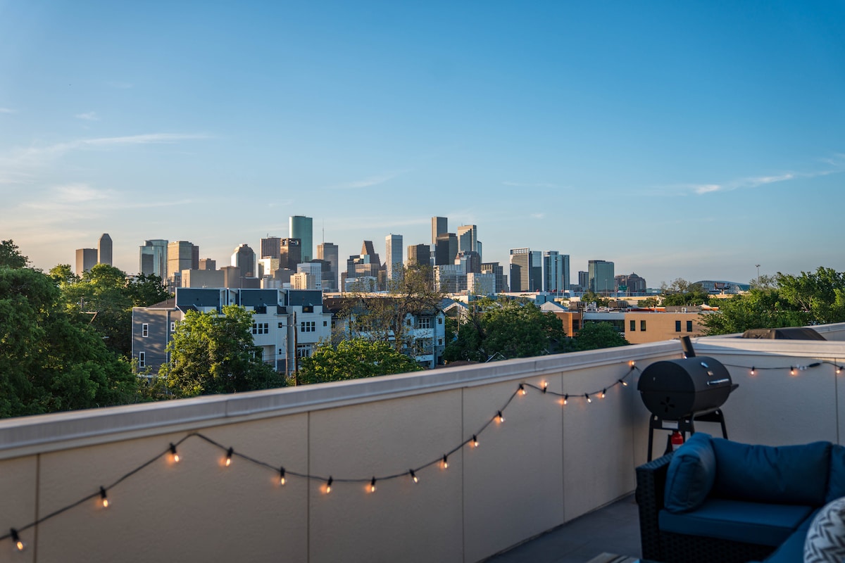 A spacious rooftop lounge offers sweeping views of the Houston skyline, framed by greenery. String lights softly illuminate the area, which includes a grill and comfortable seating for relaxation.