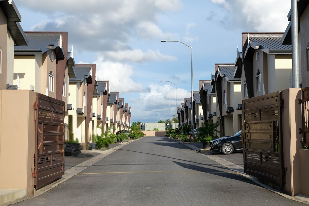 A wide view of a gated townhouse community is presented, featuring symmetrical rows of buildings with sloped roofs. The entrance showcases open, decorative gates, and a smooth asphalt road leads into the neighborhood, flanked by greenery and blue skies, creating a peaceful atmosphere.