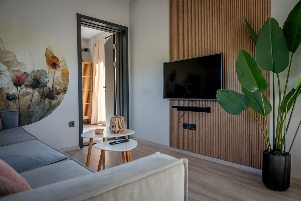 A modern living area is presented, featuring a large, light gray sectional sofa. A flat-screen television is mounted on the wall opposite, with a wood accent wall behind. A vibrant artwork depicting floral elements is displayed nearby, along with a potted plant and small side table.