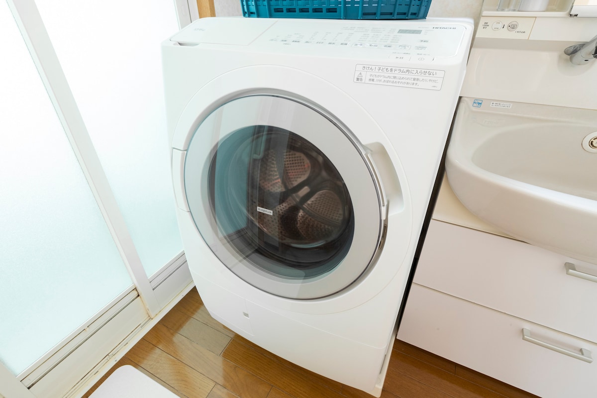 A modern washing machine with a front-loading design and a clear glass door is positioned next to a sink. The machine is white, blending seamlessly with the surrounding cabinetry. Natural light filters through a nearby window, enhancing the bright and functional space.