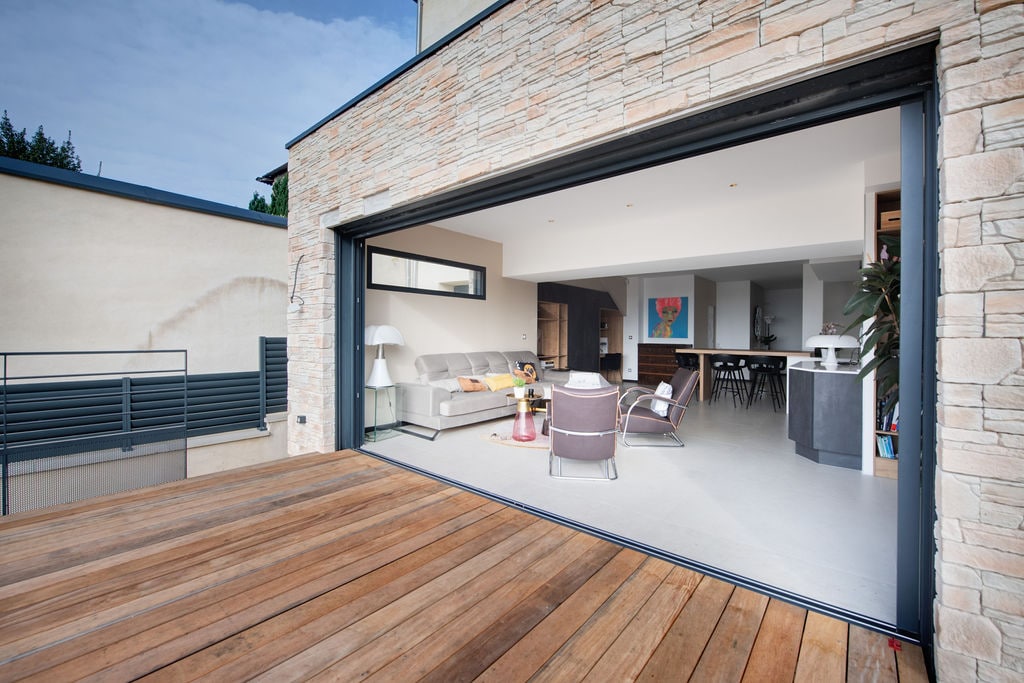 An open living area is highlighted by large sliding glass doors that connect the indoor space with an outdoor wooden terrace. The neutral-toned decor includes a comfortable seating arrangement and modern kitchen, enhancing the spacious and inviting nature of the room.