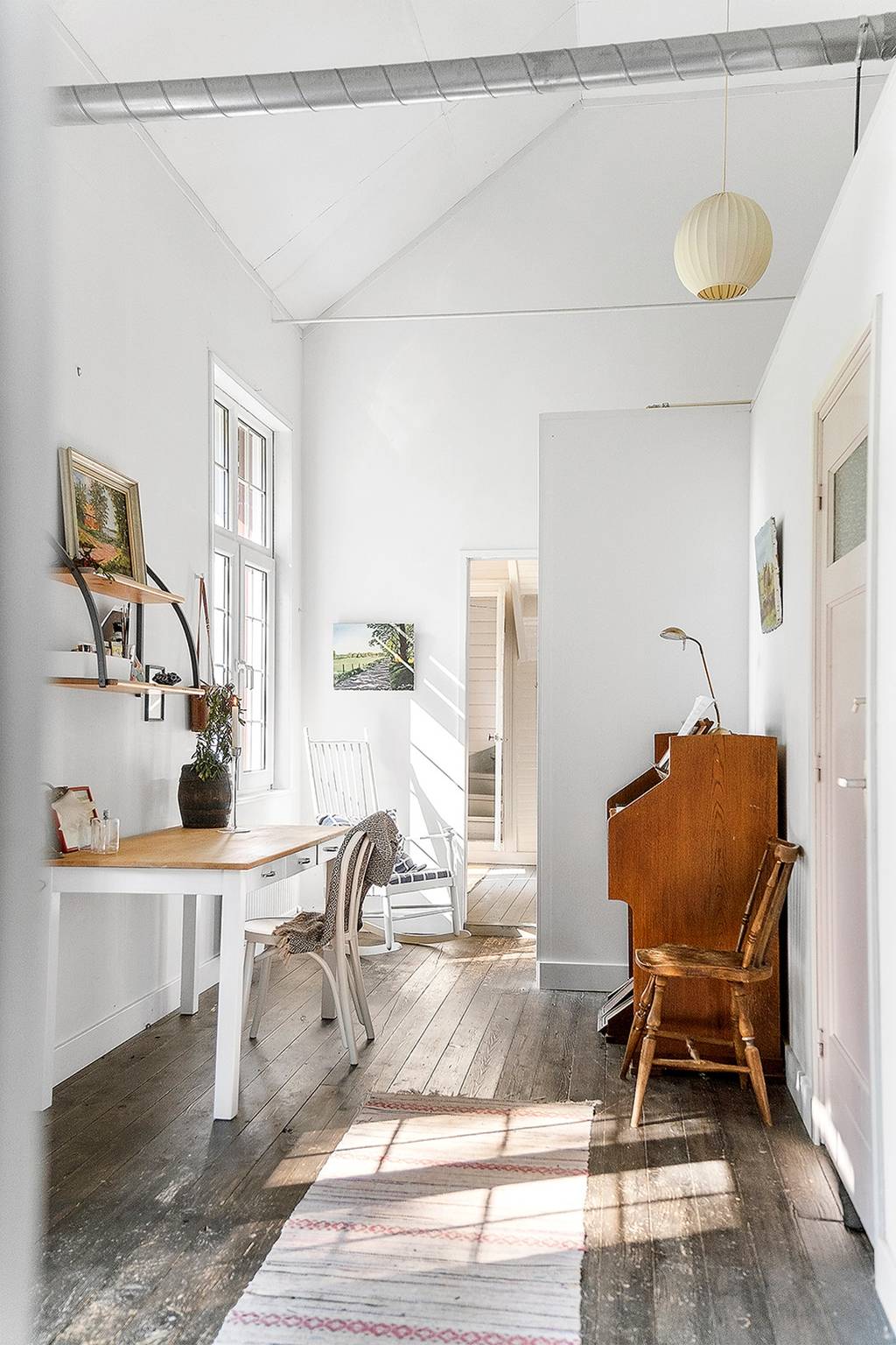A light-filled hallway is depicted, featuring white walls and wooden flooring. A small desk with a chair is positioned to one side, and a vintage wooden piano is visible. Artwork adorns the walls, while a runner adds texture to the space.