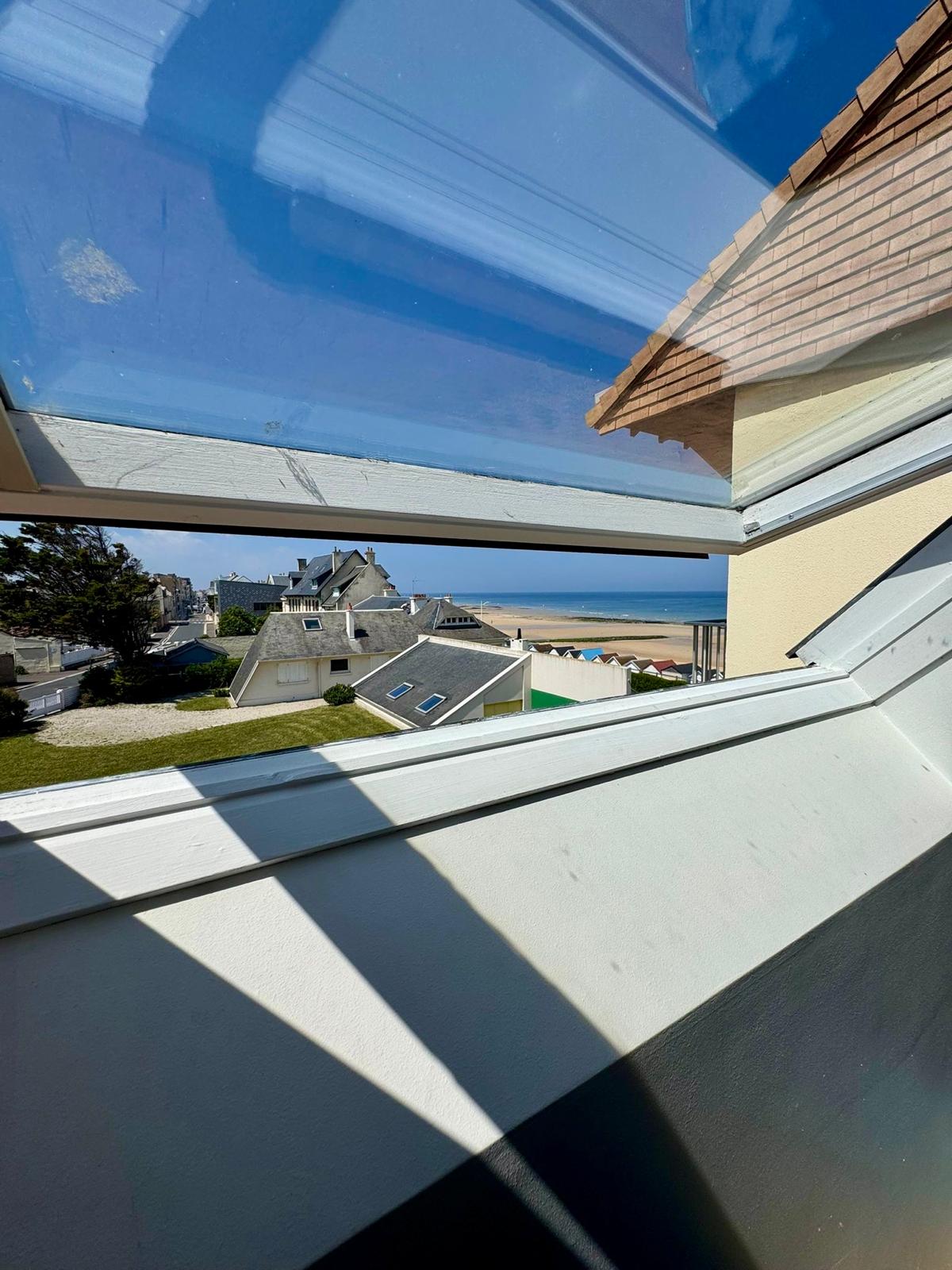 A skylight offers a view of the peaceful coastal scenery, revealing rooftops of nearby homes and the sandy beach beyond. The clear blue sky complements the serene atmosphere, allowing natural light to fill the space.