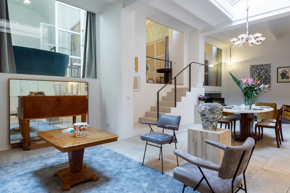 A spacious living area is illuminated by natural light streaming through skylights. Elegant modern furniture includes a round dining table, a wooden coffee table, and stylish seating. Soft-colored walls and a plush rug complement the surroundings, while a staircase provides access to the upper level.