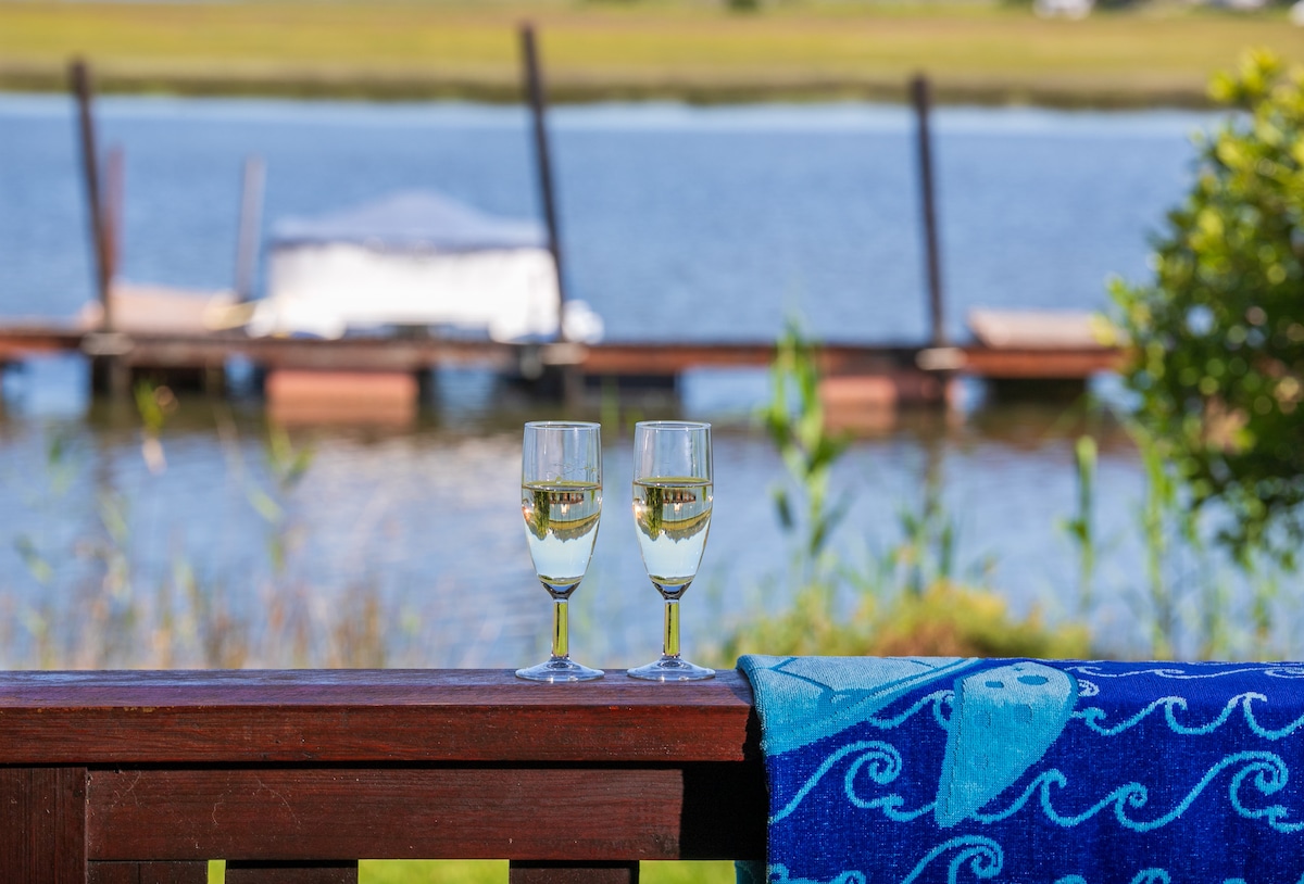 Two glasses of sparkling wine are positioned on a wooden railing, with a view of the calm water beyond. A blue towel with a wave pattern is draped over the railing. In the background, a dock is visible, accentuated by the lush green vegetation surrounding the scene.
