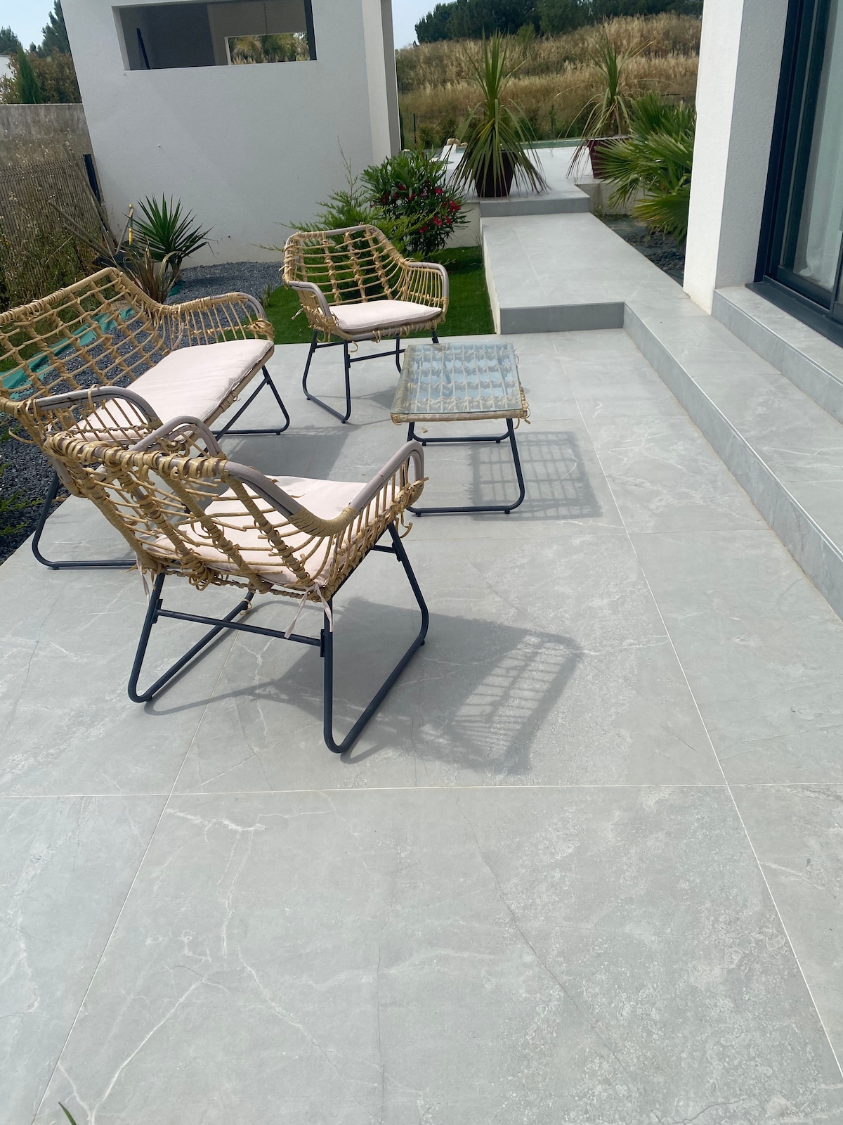 A terrace area features two rattan chairs and a small glass-top table, arranged on a light-colored stone floor. Green plants and low shrubs are visible nearby, while steps lead to the entrance of the home, enhancing the outdoor space.