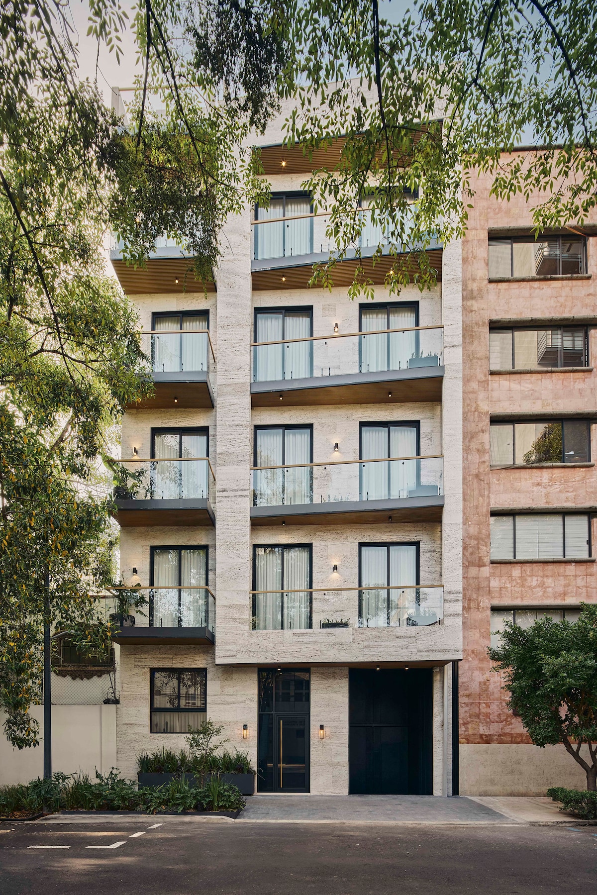 The modern building features a blend of textured stone and warm-toned materials, with multiple balconies adorned with glass railings. Lush greenery surrounds the entrance, creating a welcoming atmosphere. The façade showcases large windows that reflect natural light, enhancing the overall design.