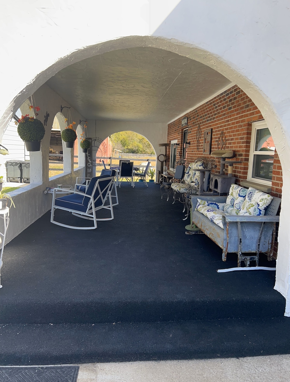 An inviting porch area is presented with arched openings and a flat black surface. Comfortable seating options include patterned cushions on benches and rocking chairs, surrounded by hanging planters. Natural light filters in, offering a welcoming space for relaxation.