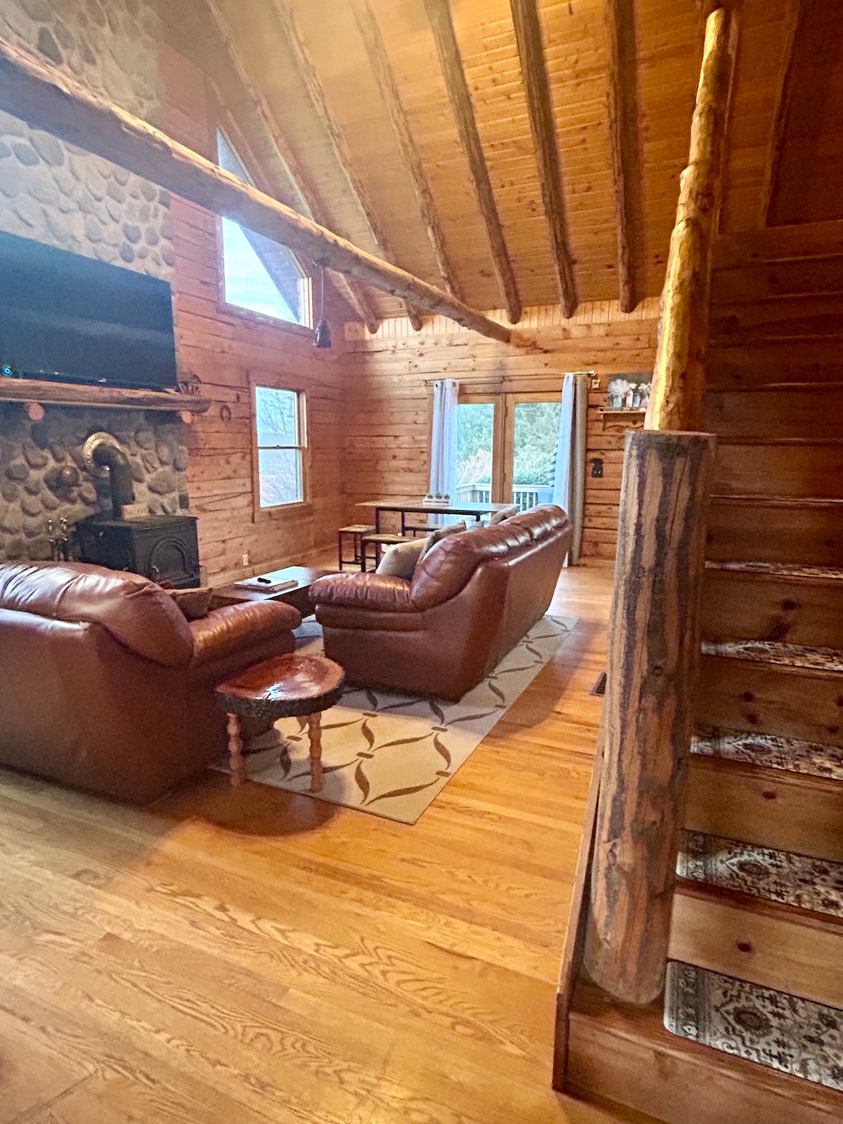The living area features a warm wood interior, with a stone fireplace as a focal point. Two leather sofas are arranged around a coffee table, complemented by a rug. Large windows allow natural light to illuminate the space, while a wooden staircase leads to the upper level.