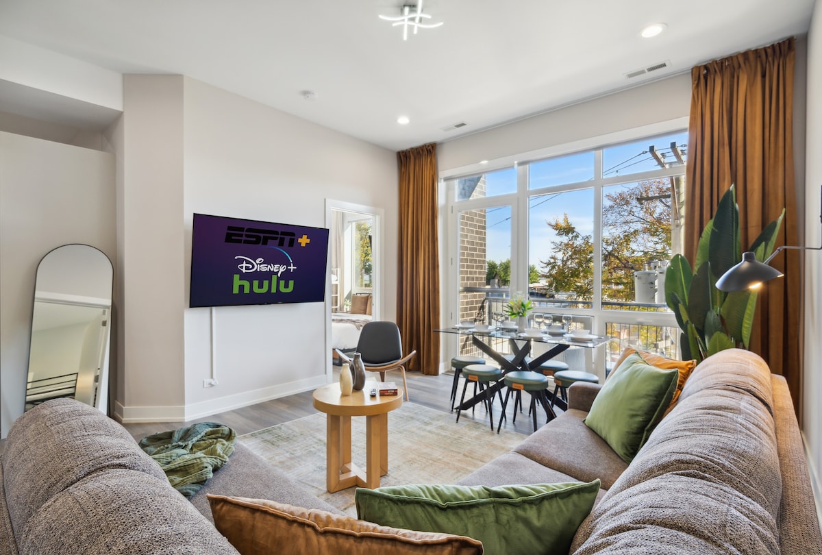 A modern living area features a large sectional couch facing a wall-mounted television displaying streaming services. Natural light floods in through expansive windows adorned with floor-length curtains. A wooden coffee table is positioned in front of a sleek dining table, complementing the neutral decor.