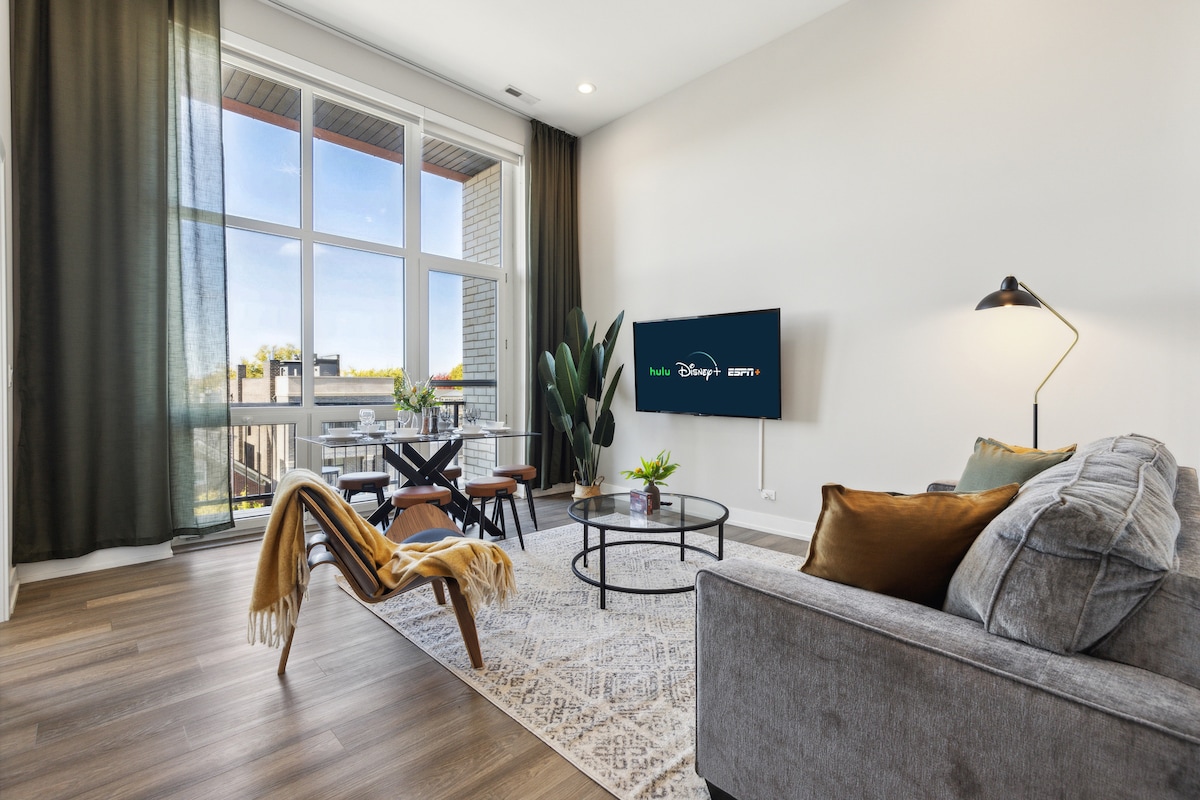 A bright living area features large windows allowing natural light to fill the space. A comfortable gray sofa is paired with a stylish armchair and a round coffee table. A flat-screen TV is mounted on the wall, while a small dining table is set near the windows.