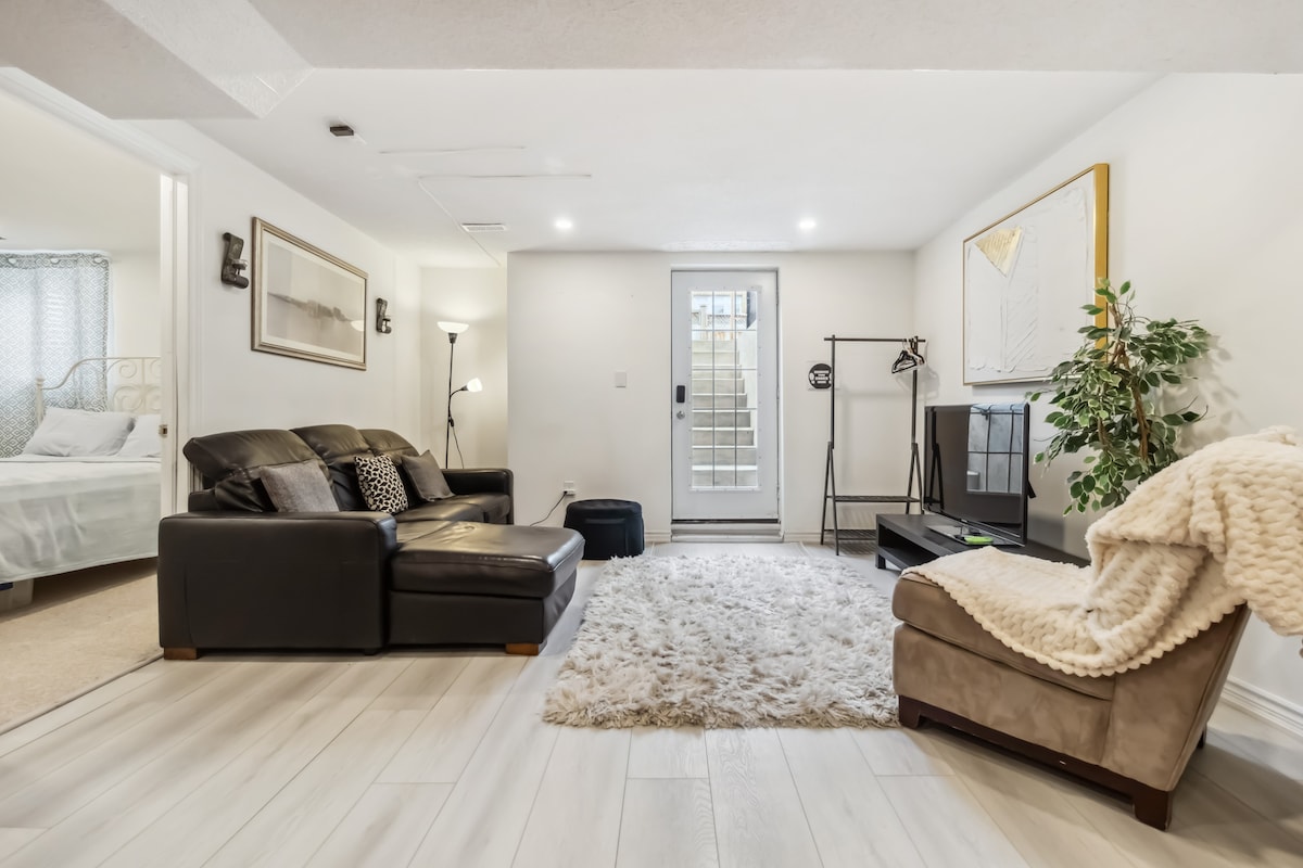 The living area features a comfortable black leather sofa positioned on a soft area rug, complemented by a cozy throw blanket. A flat-screen TV is placed on a stand, and natural light enters through the glass door leading to the outside. A chair and plant enhance the space.