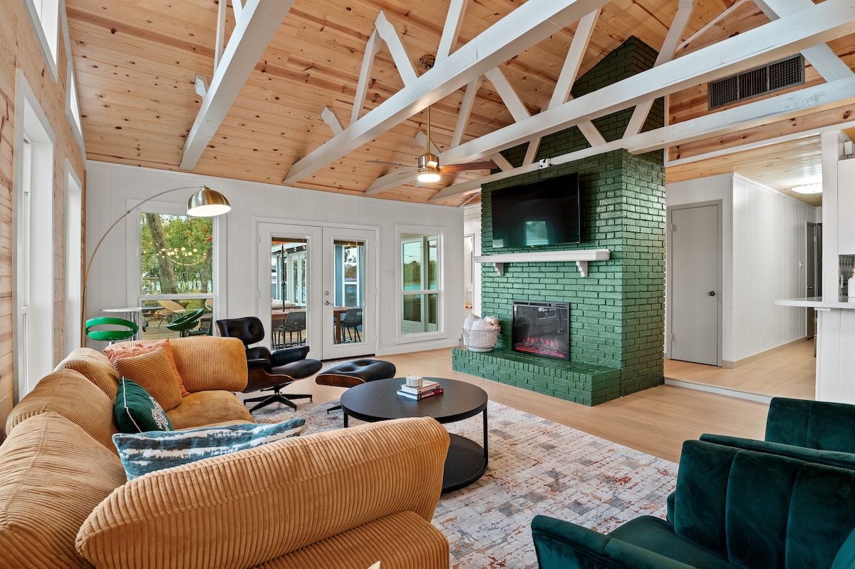 The living area features a spacious sectional couch with various cushions, paired with an accent chair. A round coffee table is centrally located, and a green brick fireplace is present. Large windows offer views of the outdoors, while exposed wooden beams enhance the ceiling.
